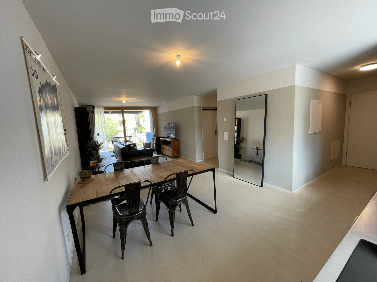 Open-plan living and dining area with a wooden dining table, metal chairs, a sofa, TV stand, and a large mirror. The space features light-colored walls and flooring, with a sliding glass door leading to a balcony.
