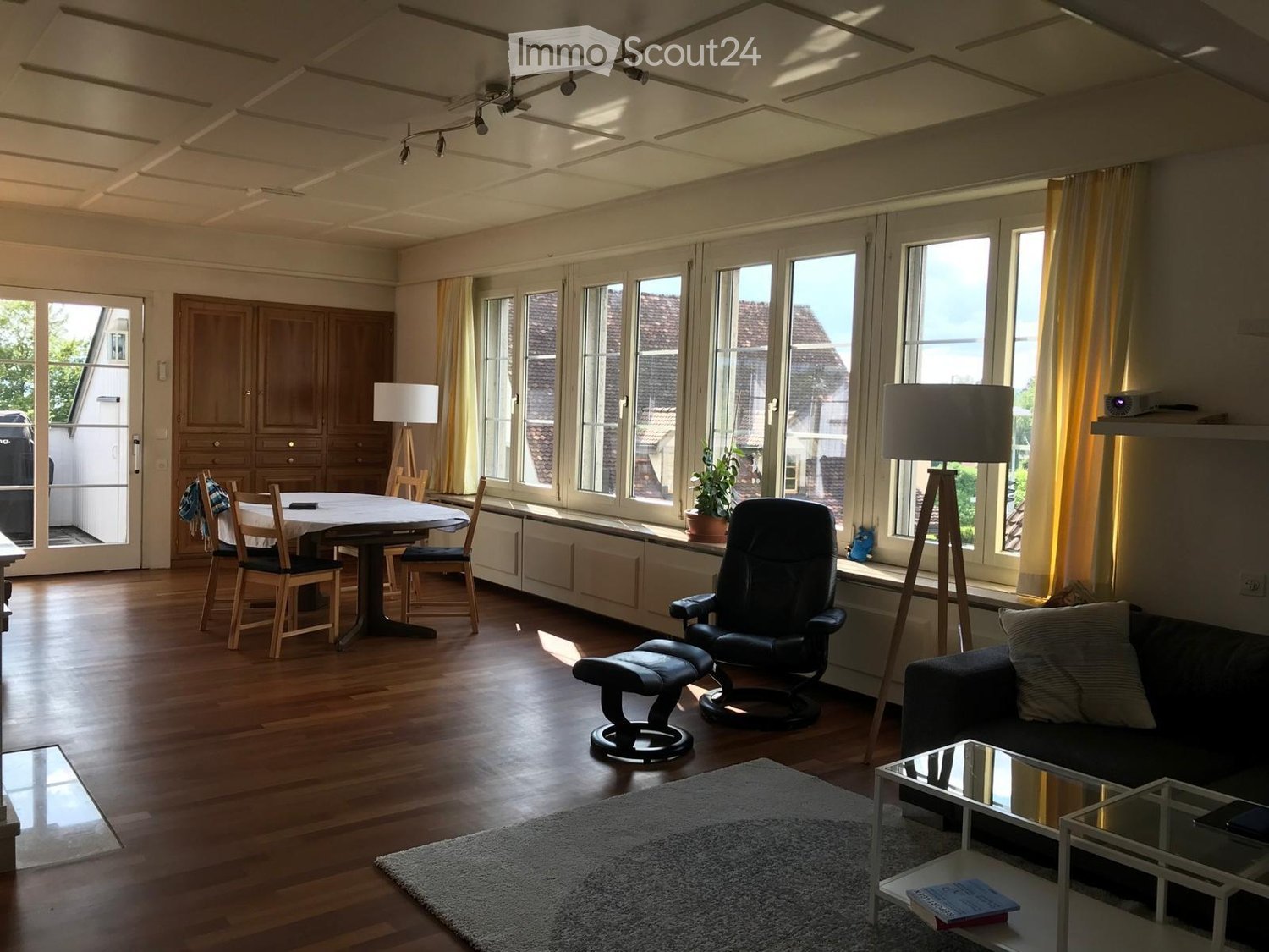 Bright living room with wooden flooring, large windows, a sofa, coffee table, dining area with table and chairs, and a cabinet.