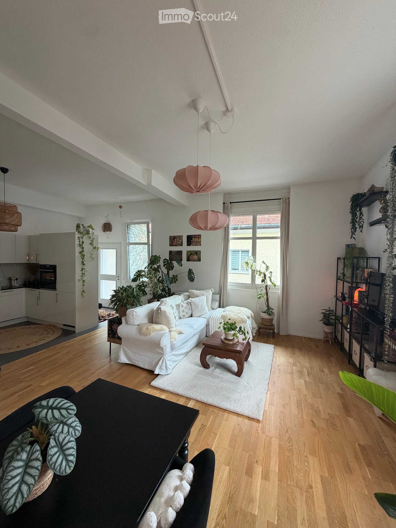 Bright living room with wooden flooring, white walls, and large windows. Features a white sofa with cushions, a small wooden coffee table, a black side table, pendant lights, and several potted plants.