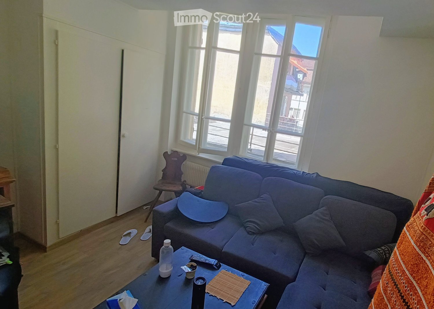 A modern living room with a blue sectional sofa, coffee table, wooden chair, and large windows offering a view of a balcony. The room features a wooden floor and white walls.