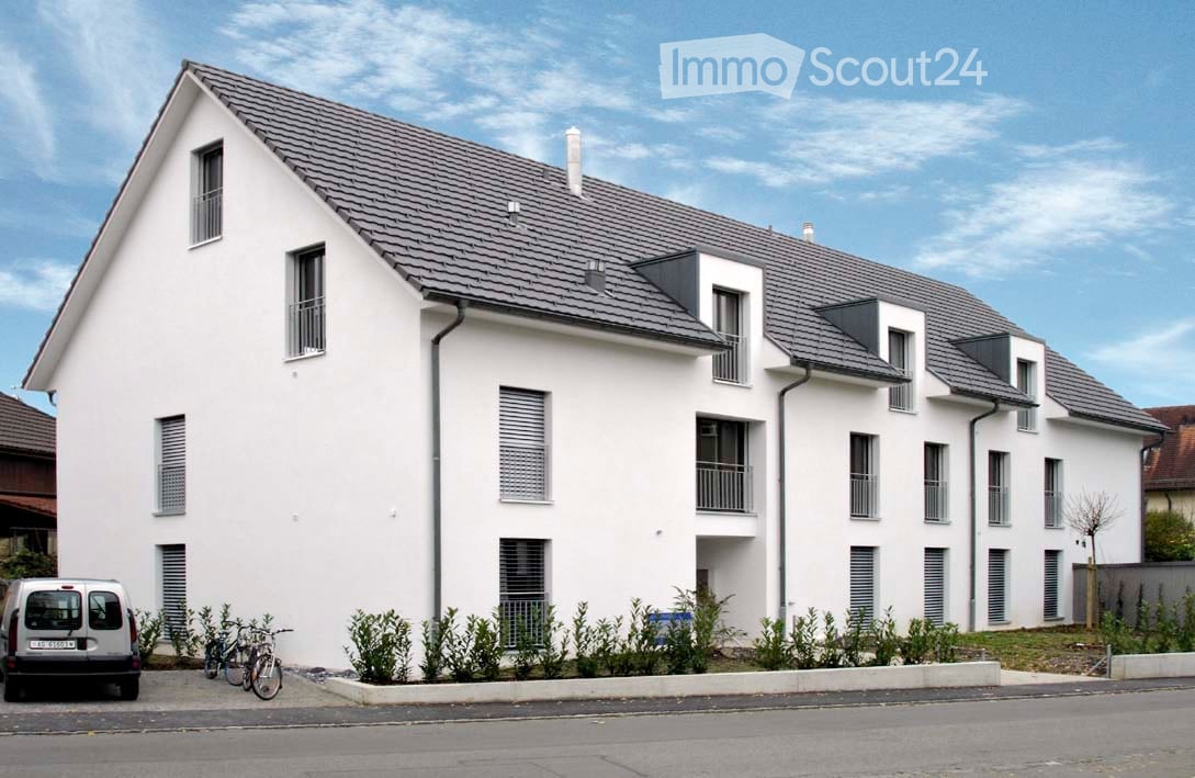 Modern two-story white building with grey roof, multiple windows, balconies, and small garden area in front.