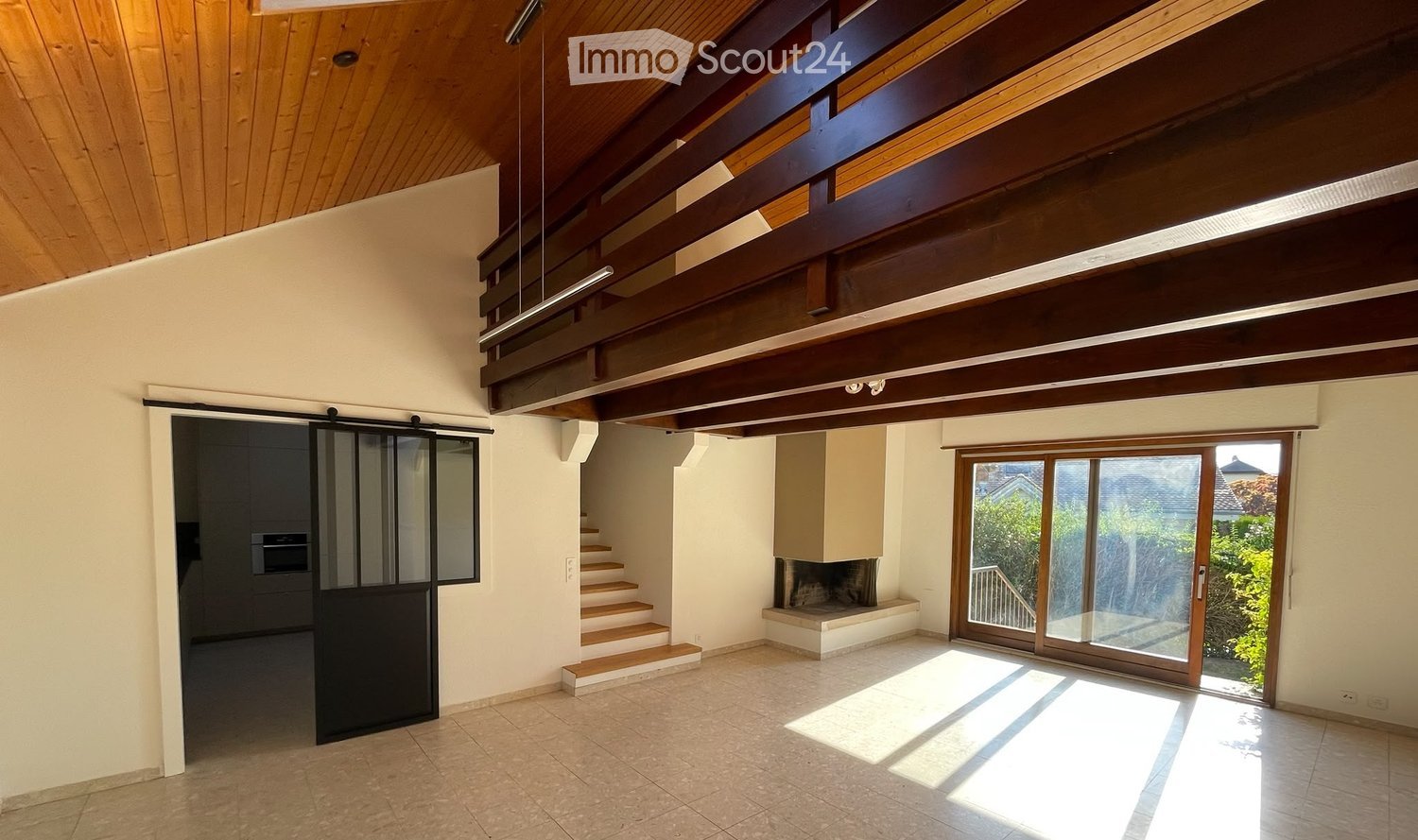 Bright, open living space with wooden ceiling beams, large sliding glass doors leading to a balcony, and a staircase leading to an upper level.