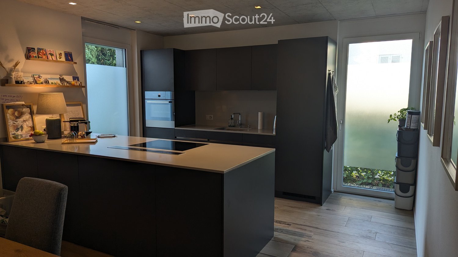 Modern kitchen with dark cabinets, a central island, built-in oven, sink, and various decorative items on shelves and counters.