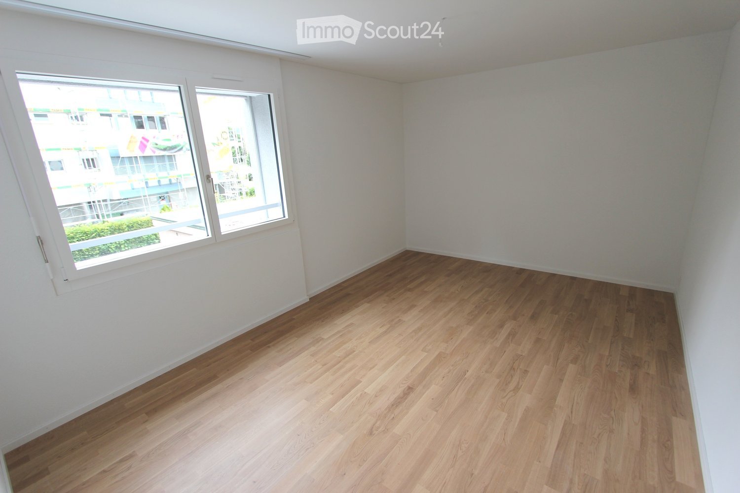 Empty room with wooden flooring and white walls, featuring large windows with a view of an outdoor area.