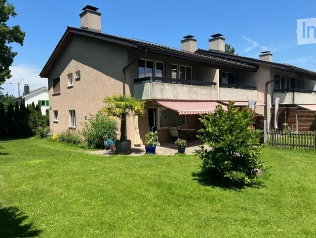 Two-story house with a balcony, surrounded by a well-maintained lawn and garden area with potted plants and outdoor furniture.