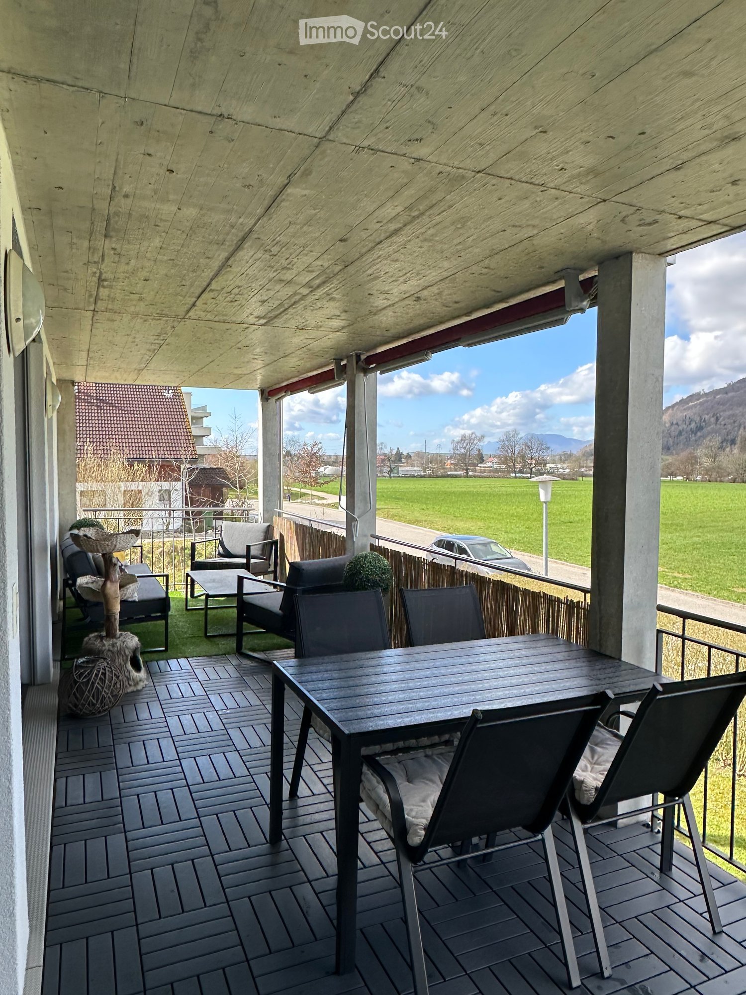 Balcony with a view of a green field and mountains, featuring a wooden table, chairs, a sofa, and a cat tree.