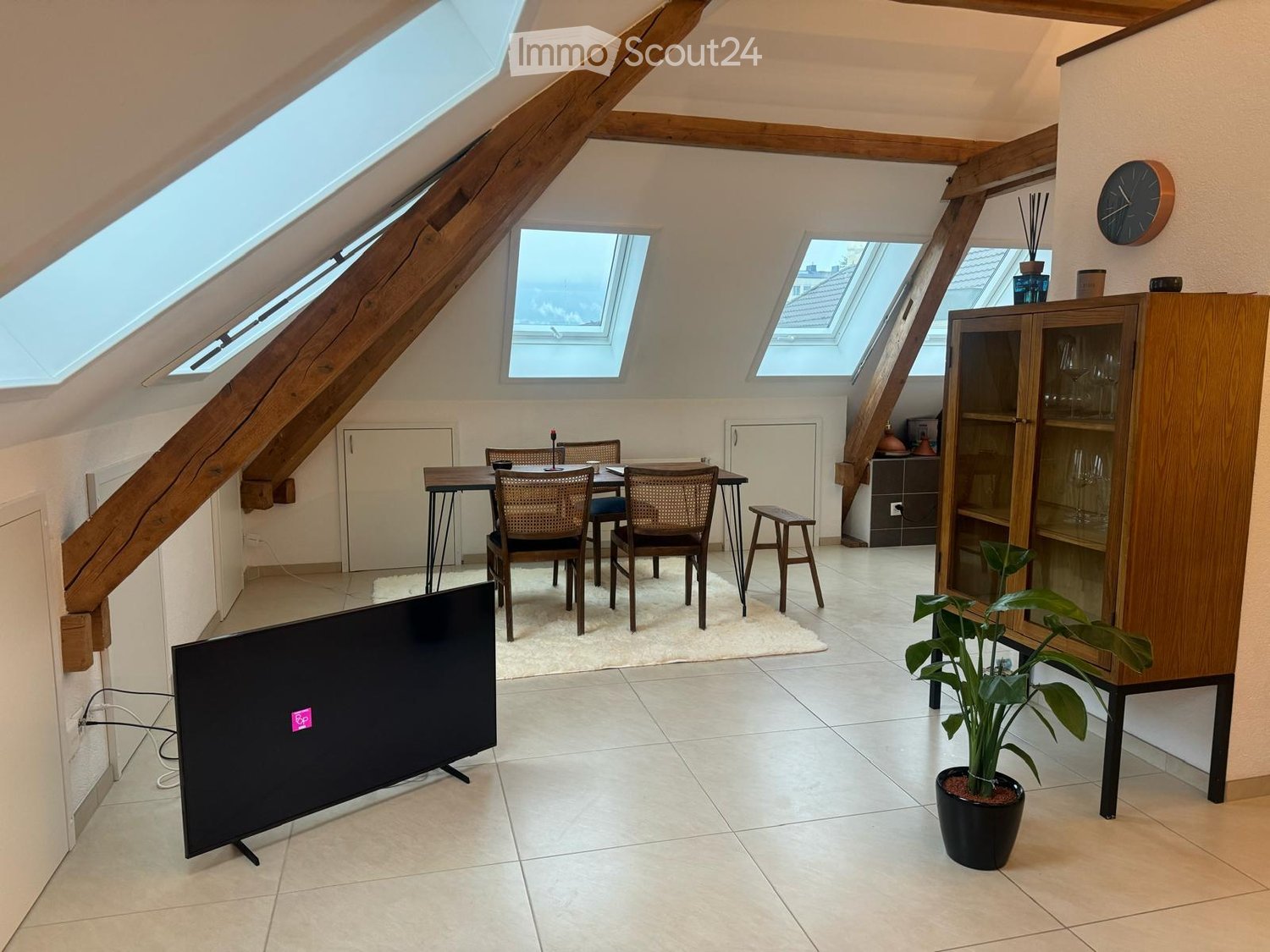 Bright attic room with skylights, tiled floor, television, dining table with chairs, cabinet, and potted plant.