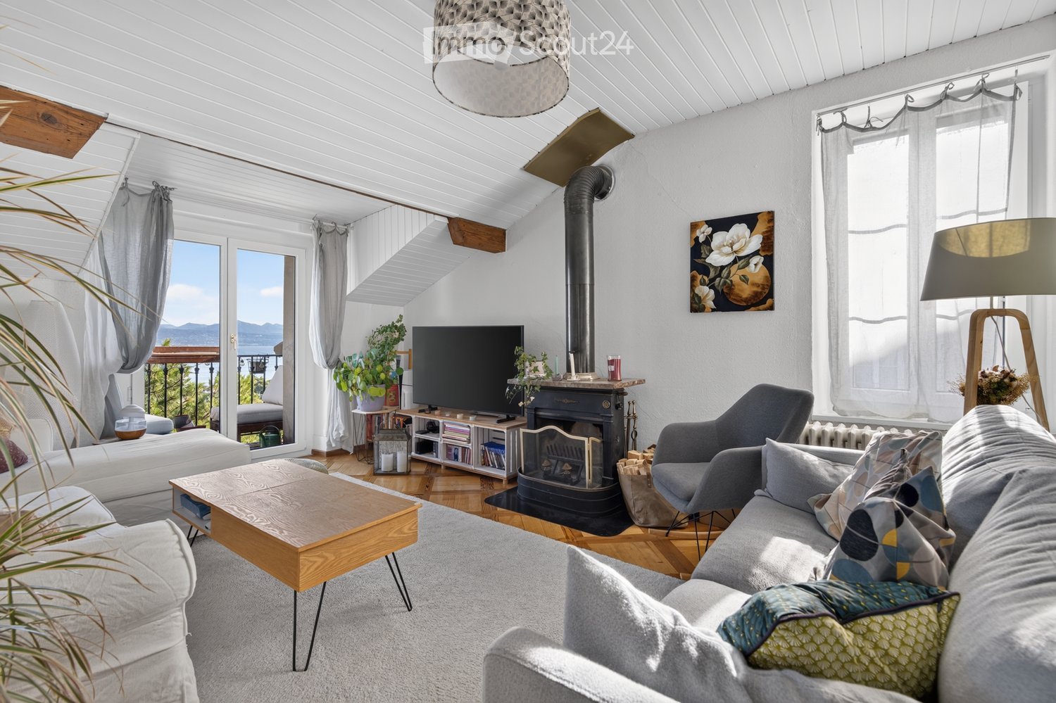 Bright living room with white walls, wooden floors, and a skylight ceiling. Features a grey sofa, wooden coffee table, armchair, TV, fireplace, and decorative plants.