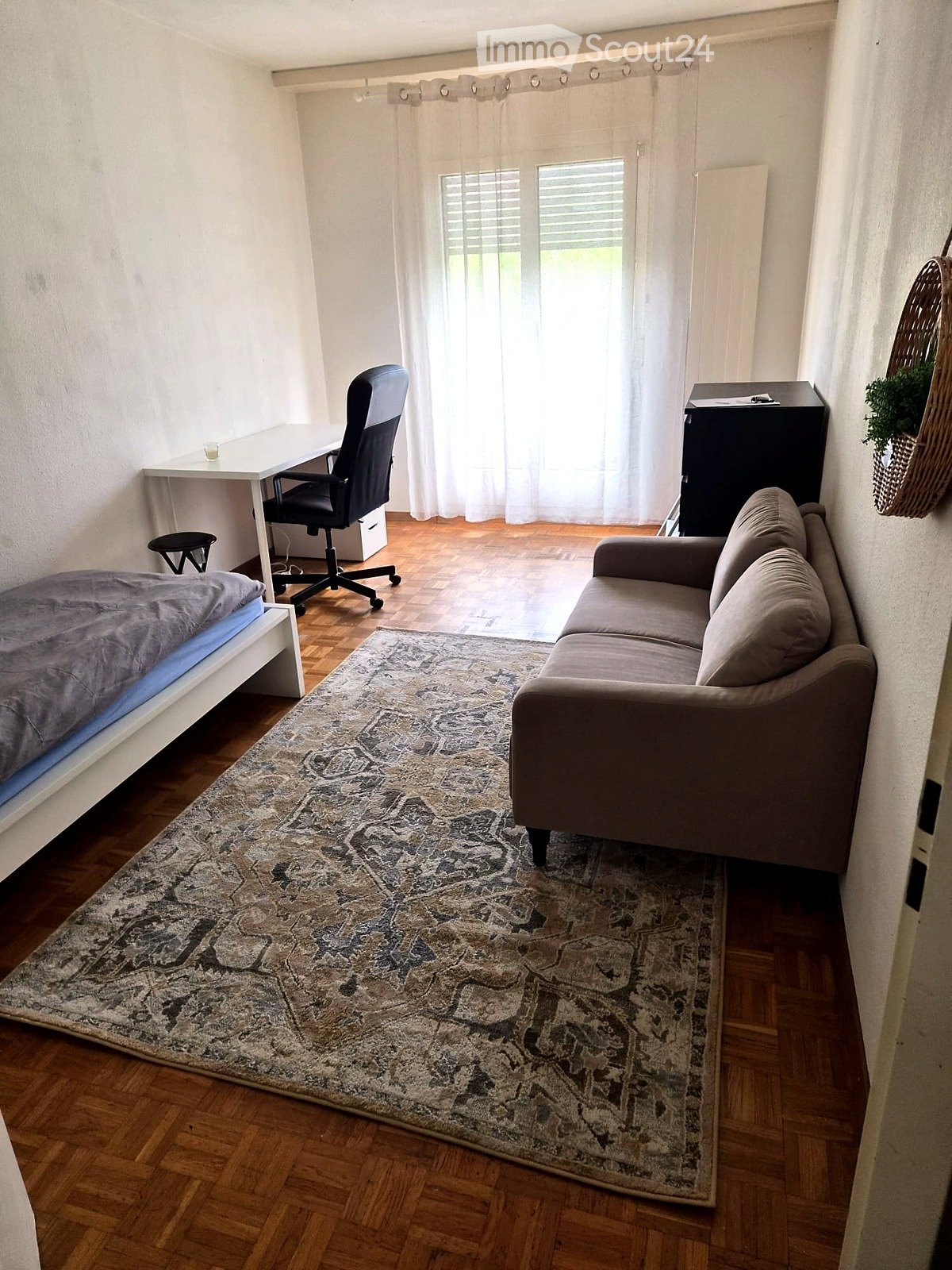 A compact, well-furnished room with a bed, sofa, desk, and chair. The room features wooden flooring, a patterned rug, and large windows with sheer curtains.