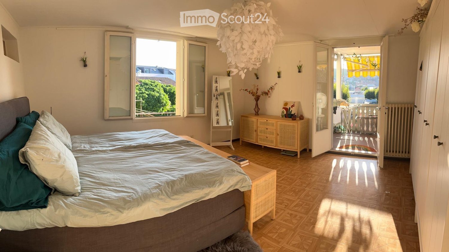 Bright bedroom with wooden flooring, large windows, a bed with pillows and blankets, a nightstand, a dresser, a mirror, and a balcony access.