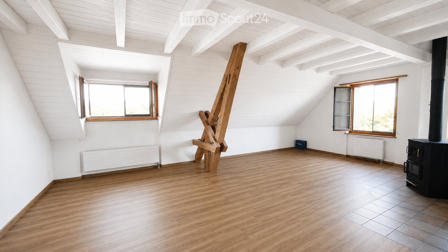 Empty attic room with wooden floors, white walls, exposed wooden beams, and two windows. A black wood-burning stove is present, but no furniture or furnishings.