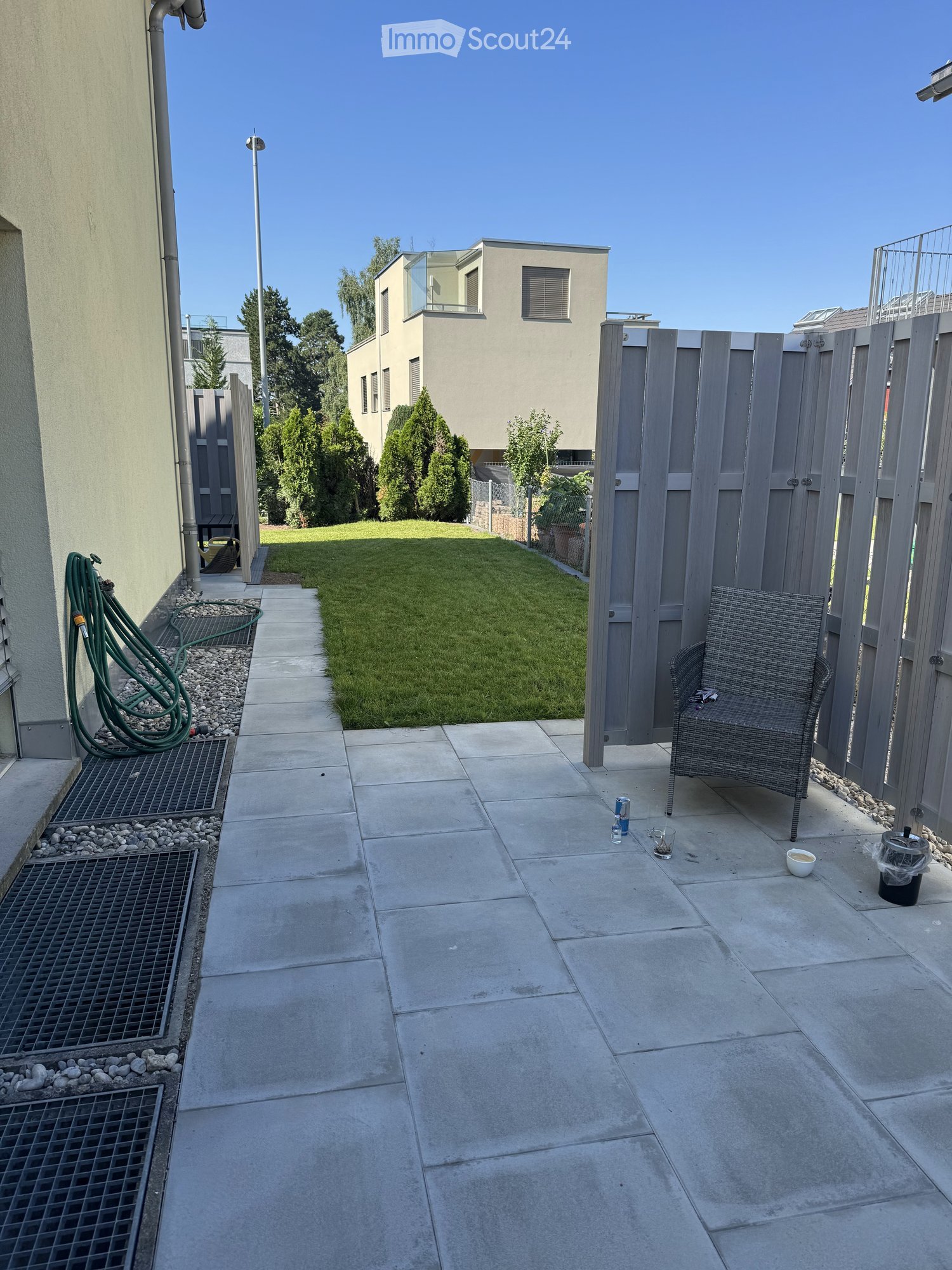 Patio area with concrete tiles, a small grassy lawn, a grey wooden fence, and a wicker chair.