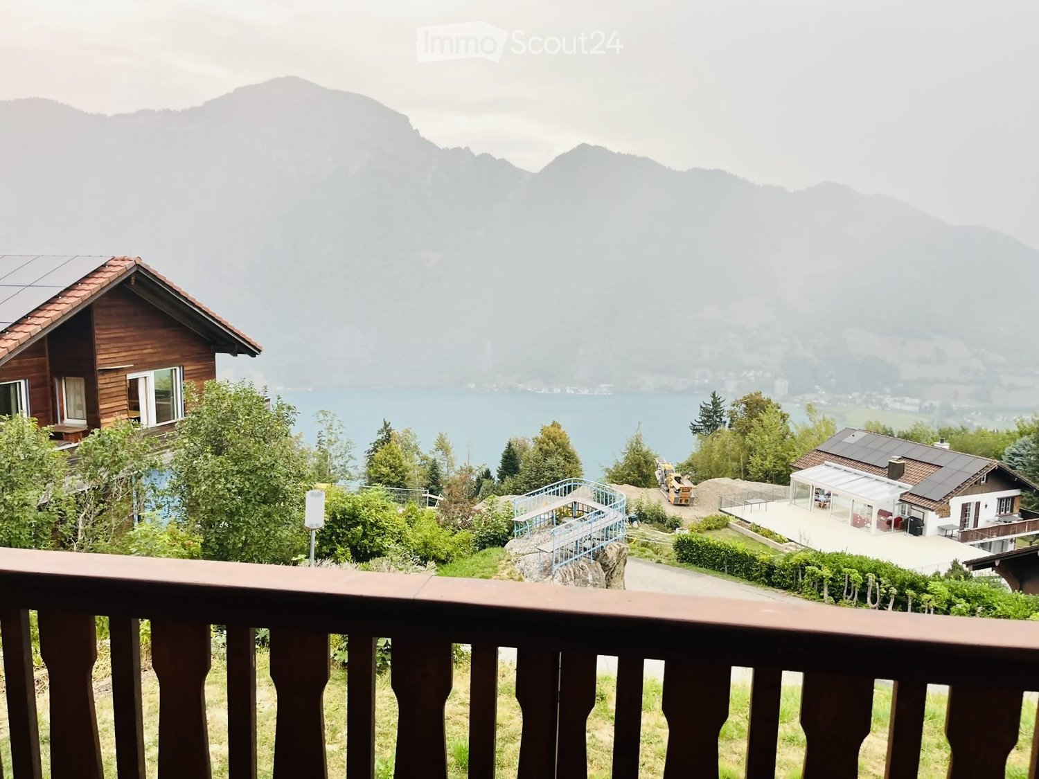 View from a balcony overlooking a scenic landscape with a lake, mountains, and several houses surrounded by greenery.