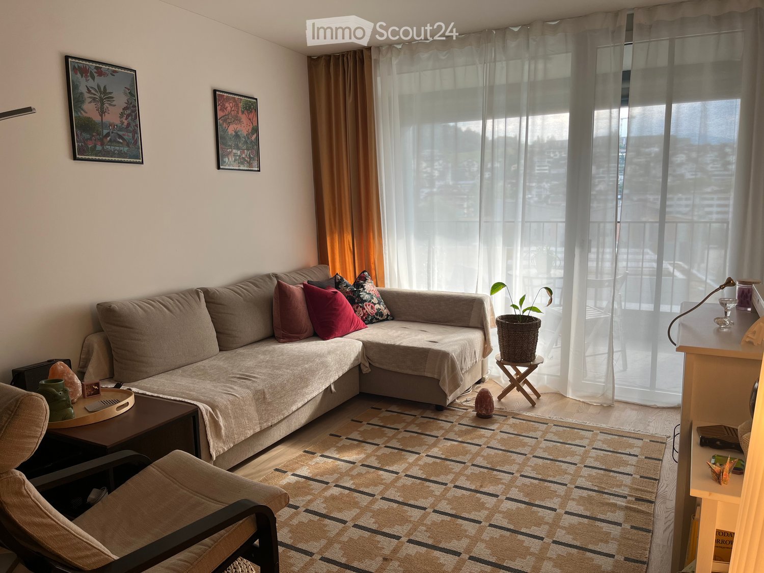 Living room with a grey sectional sofa, coffee table, armchair, potted plant, and artwork on the walls. Large windows with sheer curtains offer a view of the city.
