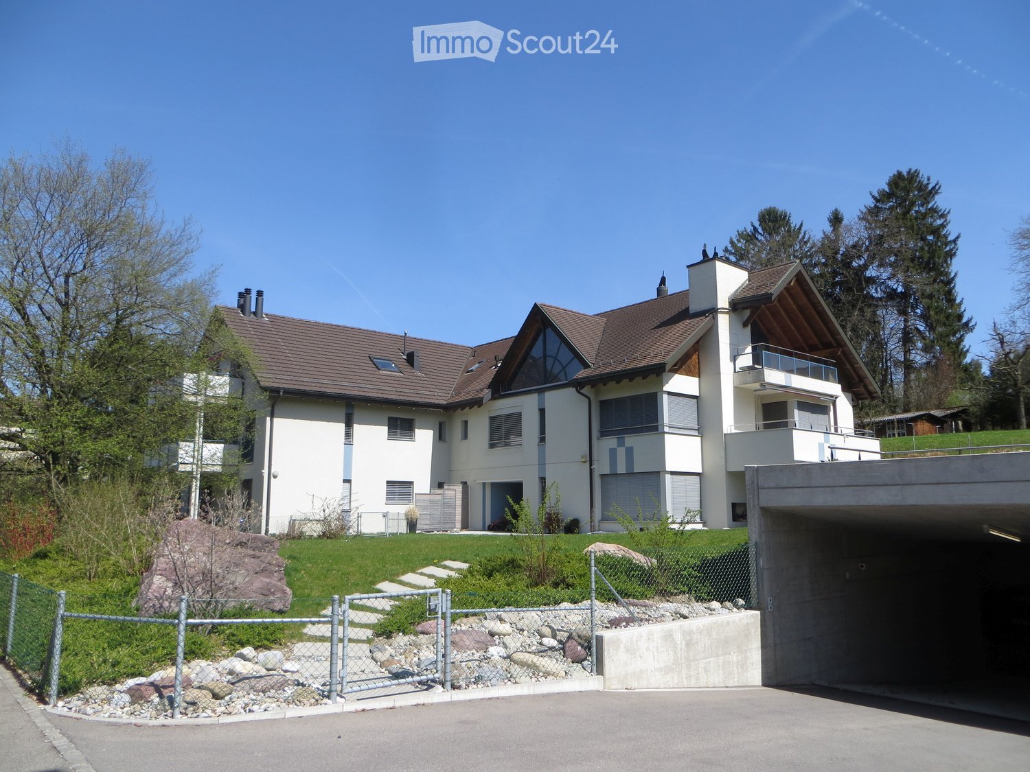 Modern two-story house with white walls, brown roof, and balcony on the upper level. The house is surrounded by a well-maintained lawn, trees, and a concrete driveway leading to a garage.