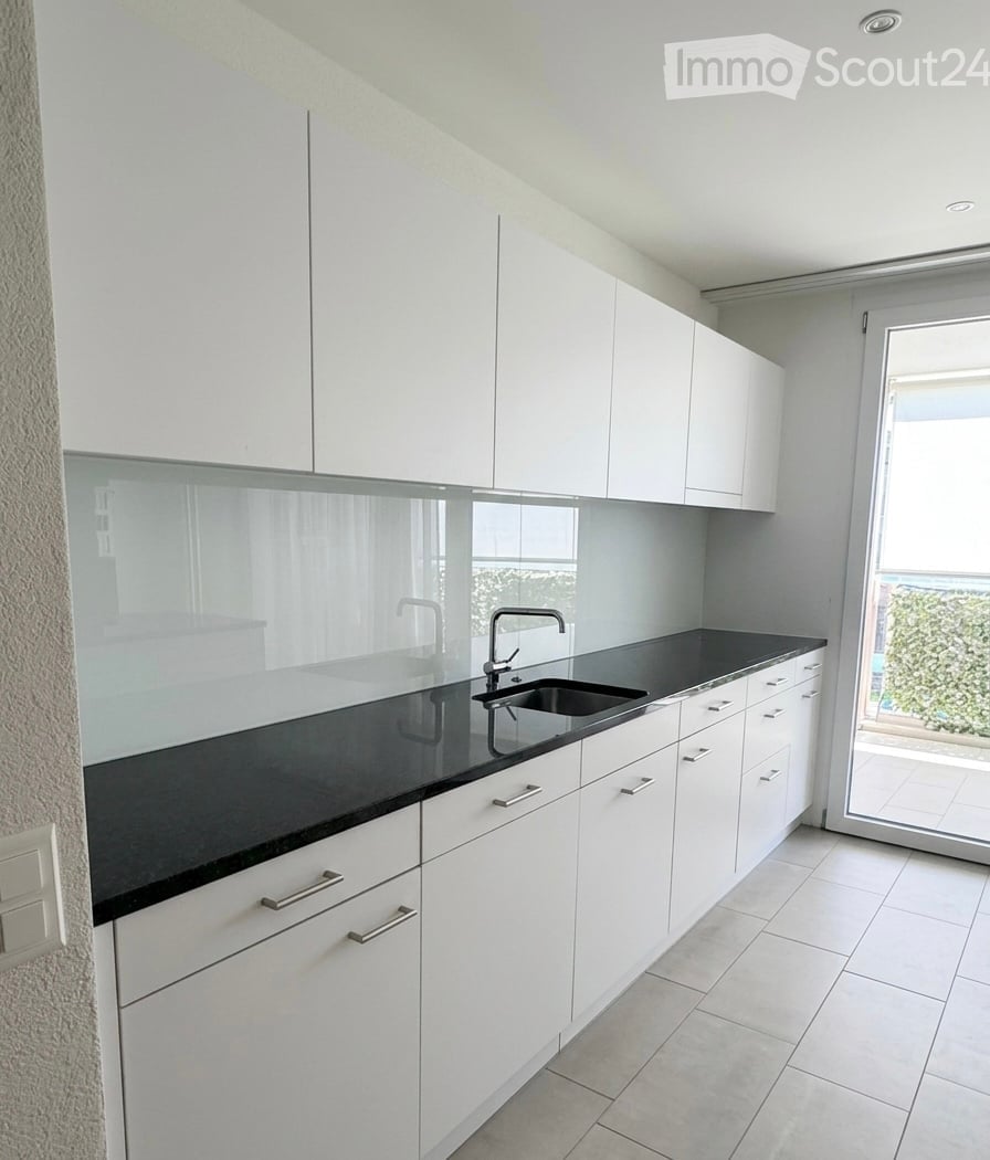 Modern kitchen with white cabinets, black countertop, and a sink with a modern faucet. The space is bright with large windows and a sliding glass door leading to a balcony.