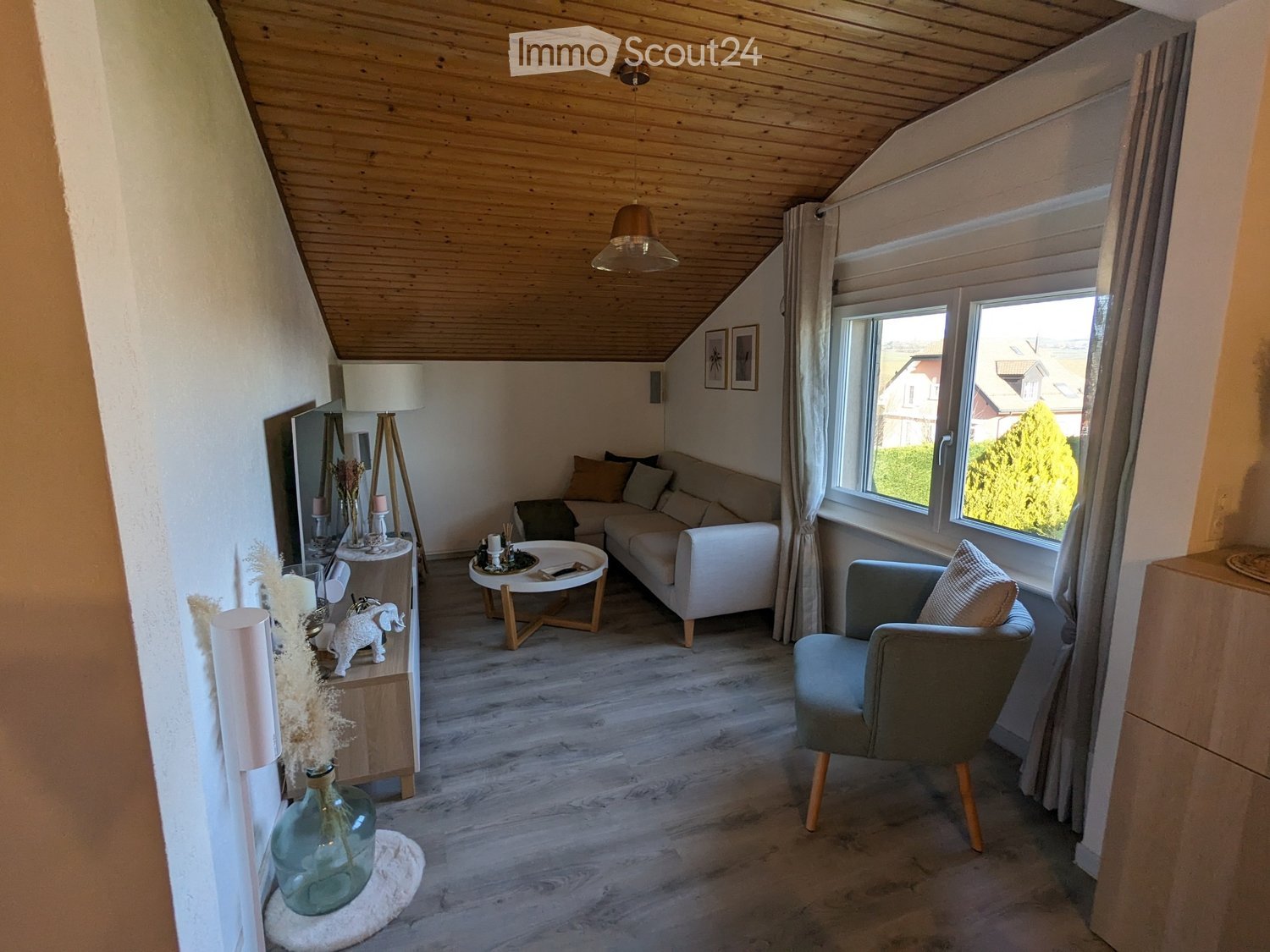 Modern living room with wooden floors, white walls, and large windows. Features a white sofa, coffee table, armchair, and decorative items including a vase and picture frames.