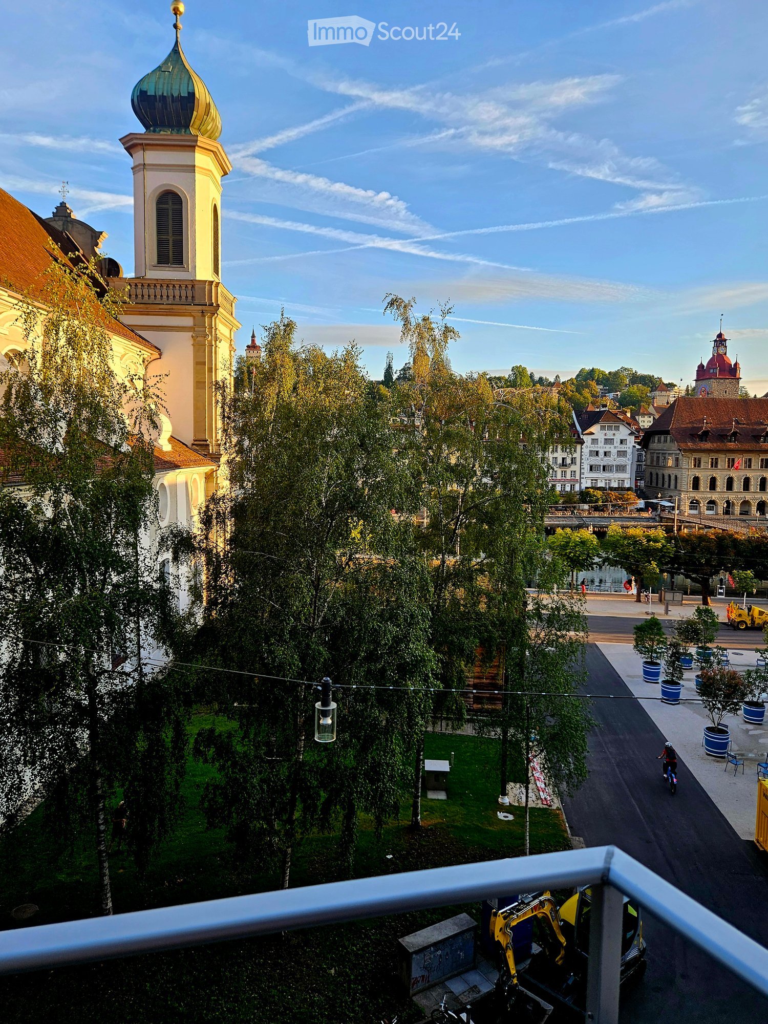 Balcony overlooking a city square with historic buildings, trees, and a small park area.