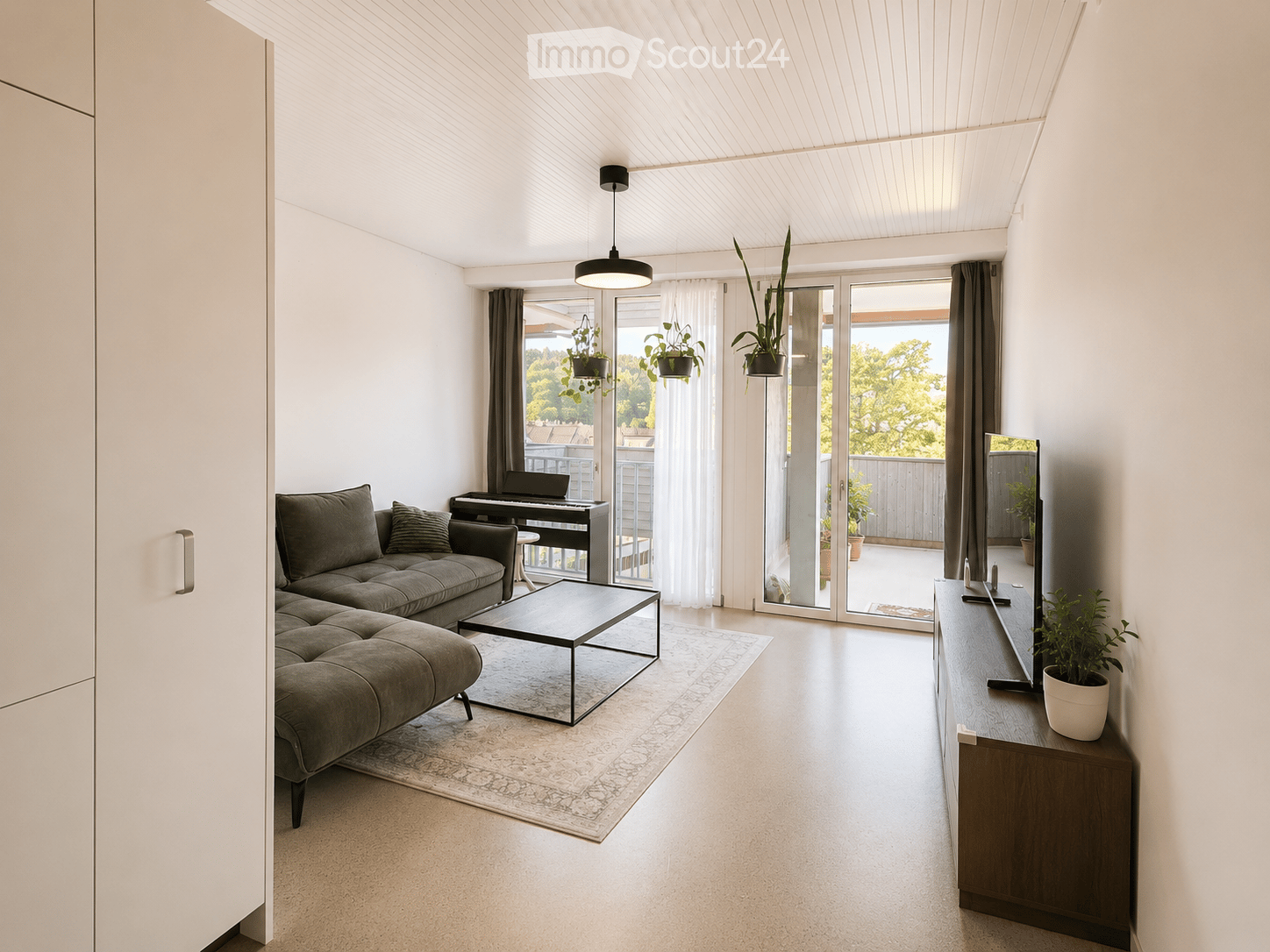 Modern living room with grey sectional sofa, coffee table, piano, TV stand, and potted plants. Large windows and sliding glass doors lead to a balcony.