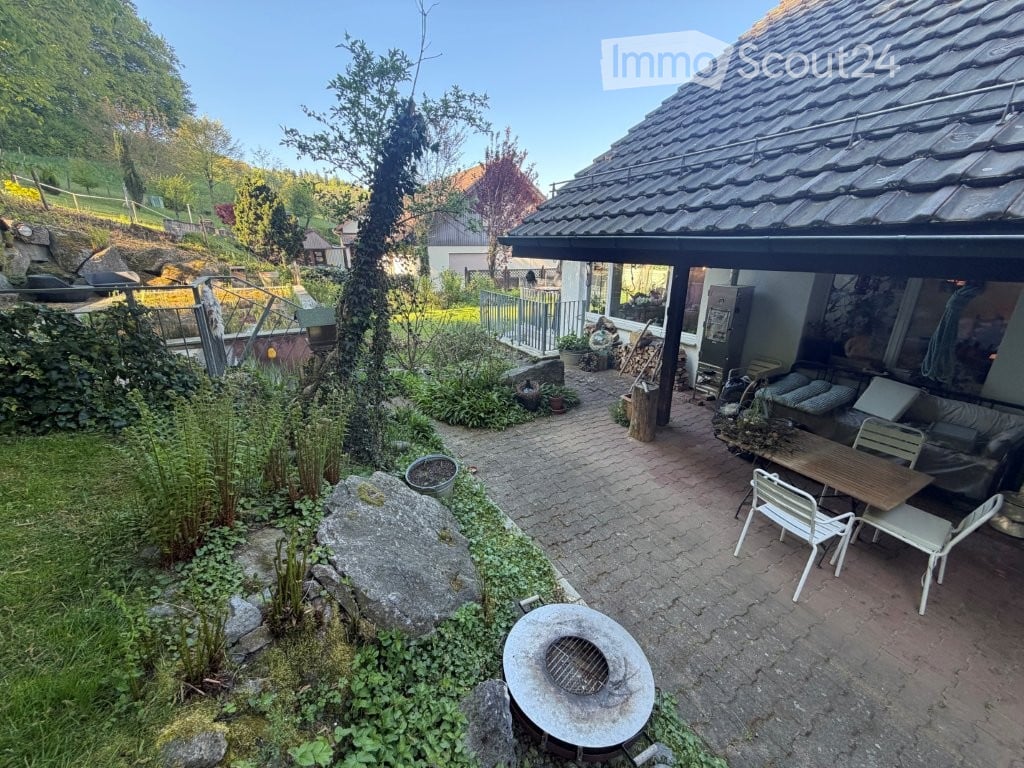 A well-maintained garden area with a patio, outdoor furniture, and a stone fire pit. The space includes a variety of plants, a small wooden table, and chairs, surrounded by a brick pathway and a wooden pergola.