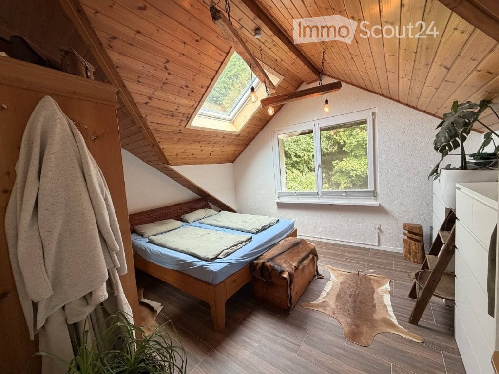 Cozy attic bedroom with wooden slanted ceilings, skylight, large window, double bed, wardrobe, and a small plant.