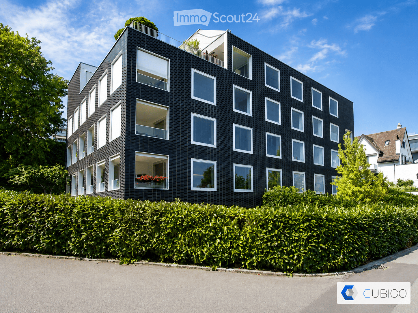 Modern black brick building with multiple large windows and balconies, surrounded by greenery and a hedge.