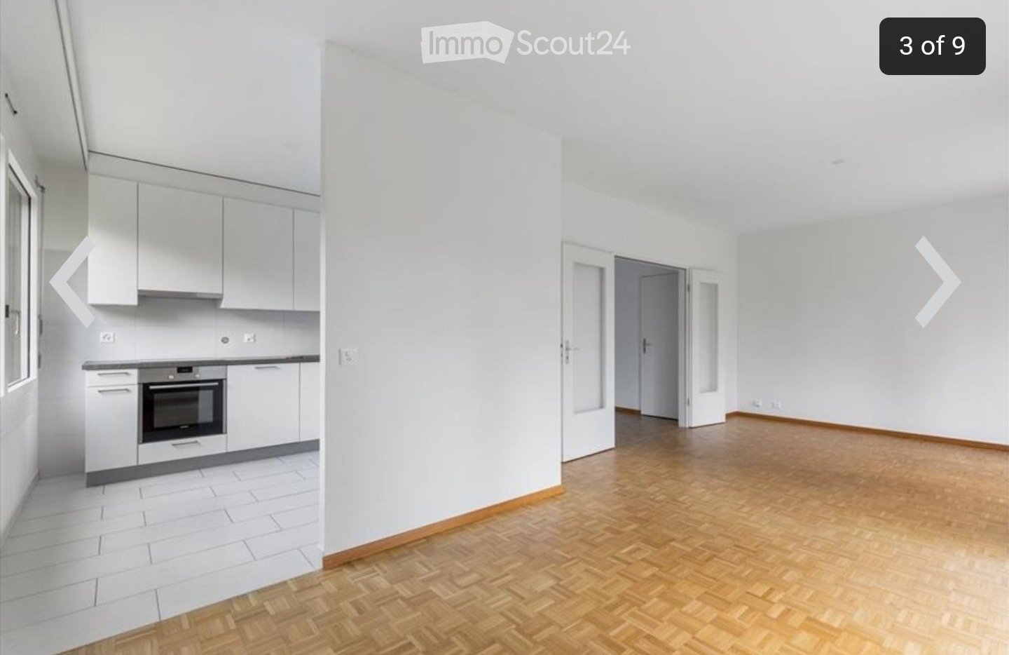 Empty room with white walls, wooden flooring, and a doorway leading to another room. The adjacent kitchen area features white cabinets and a built-in oven.