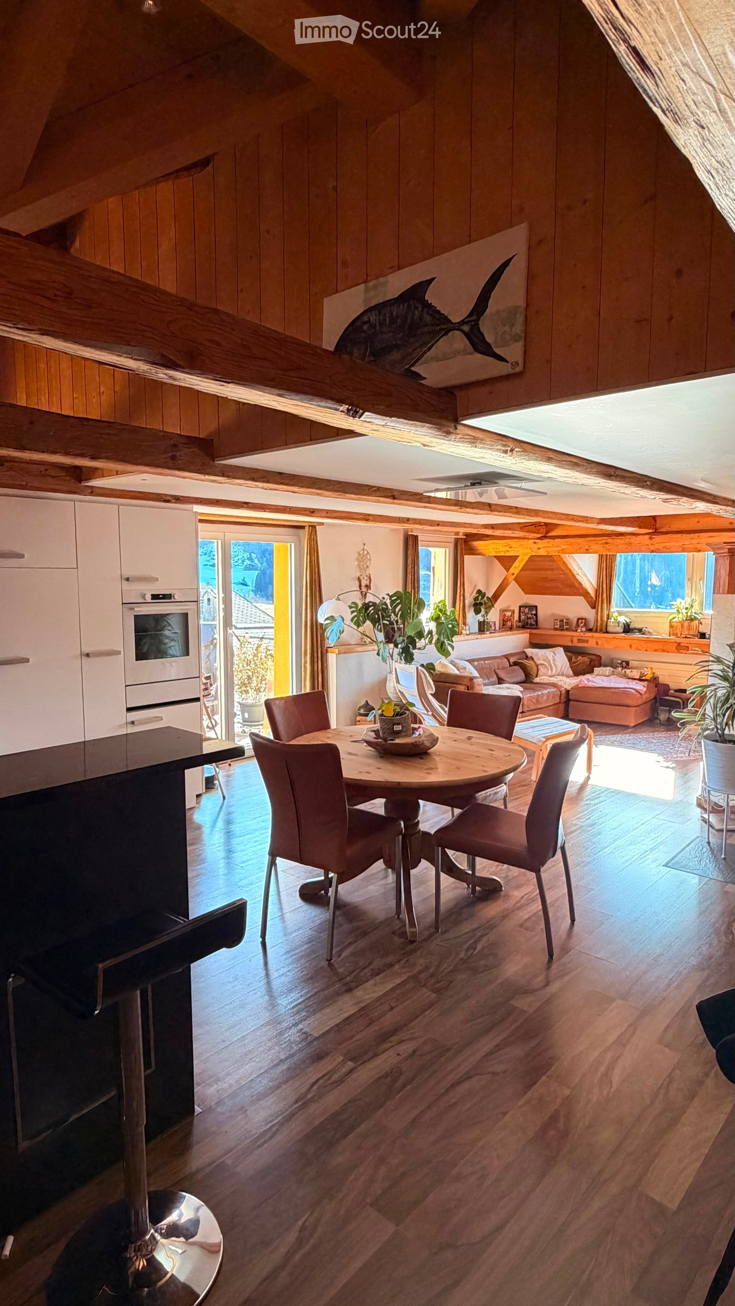 Open-plan living and dining area with wooden floors, exposed wooden beams, a round dining table with six chairs, a sofa with an ottoman, kitchen cabinets, and a modern oven. Large windows and a sliding glass door lead to a balcony.