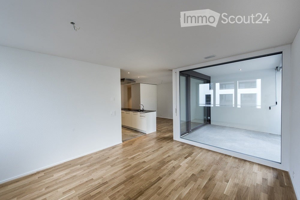 Open-plan unfurnished room with light wooden flooring, white walls, and a large glass sliding door leading to a balcony or terrace area. A small kitchenette is visible in the background.