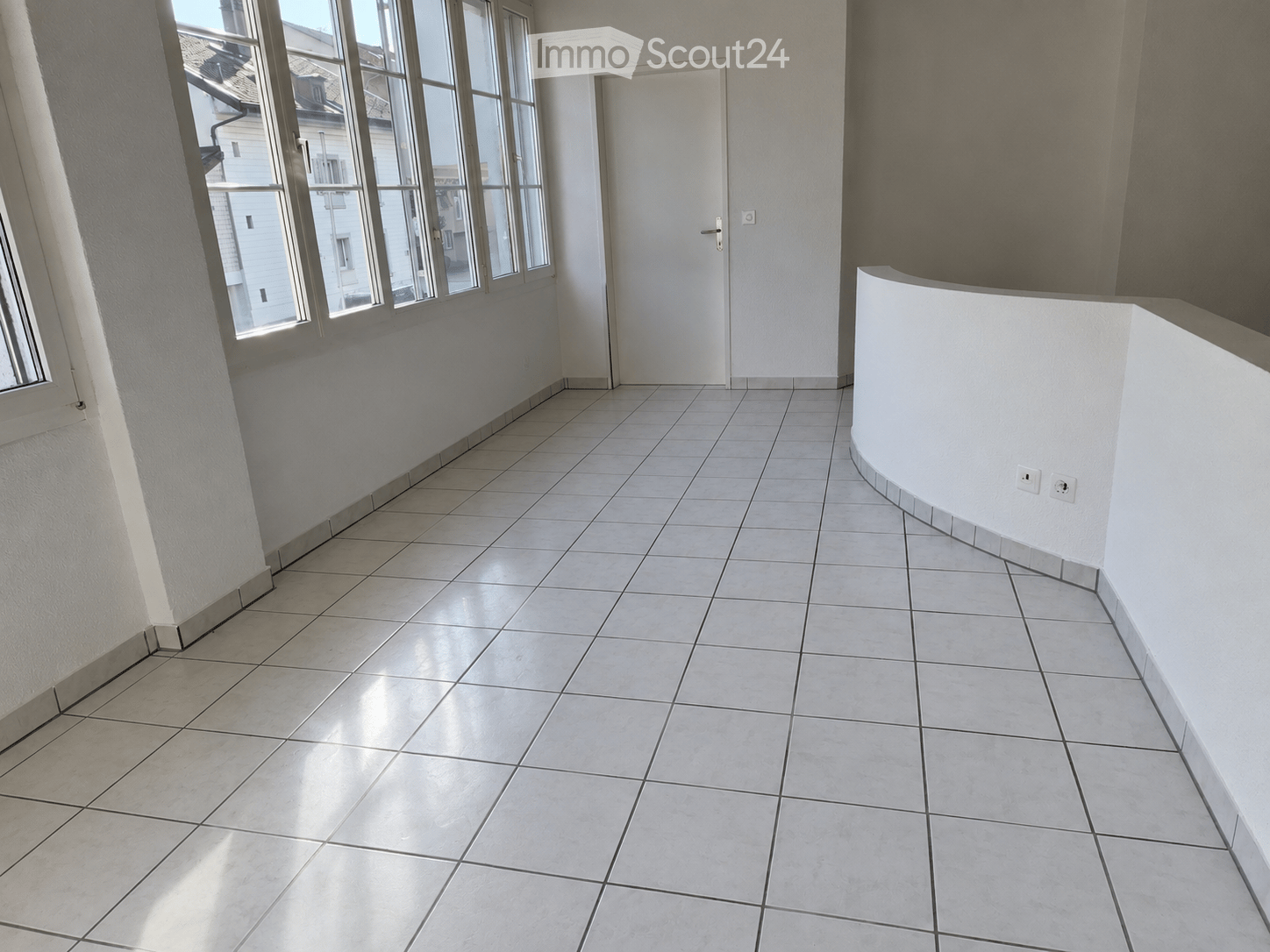 Empty room with tiled floor, white walls, large windows, and a white door.