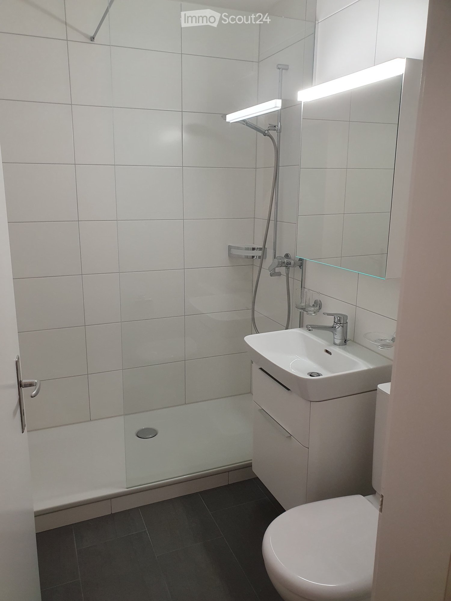 Modern bathroom with white tiled walls, a glass-enclosed shower, a white sink with a modern faucet, a toilet, and a large mirror with built-in lighting.