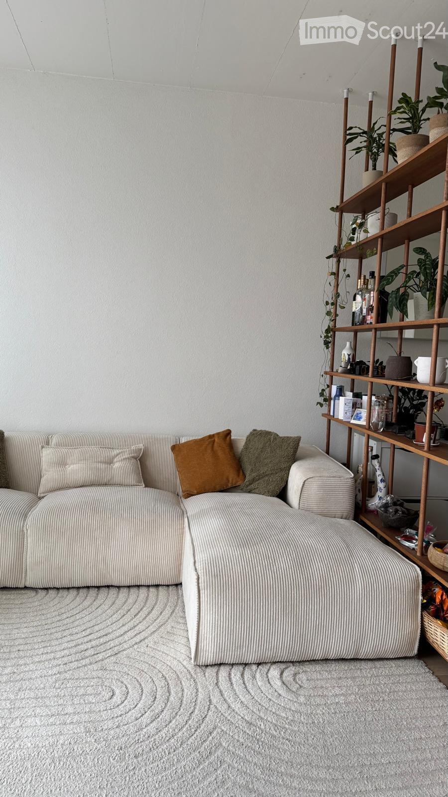 Modern living room with a white textured sofa, decorative pillows, a wooden shelf with plants and bottles, and a patterned white rug.