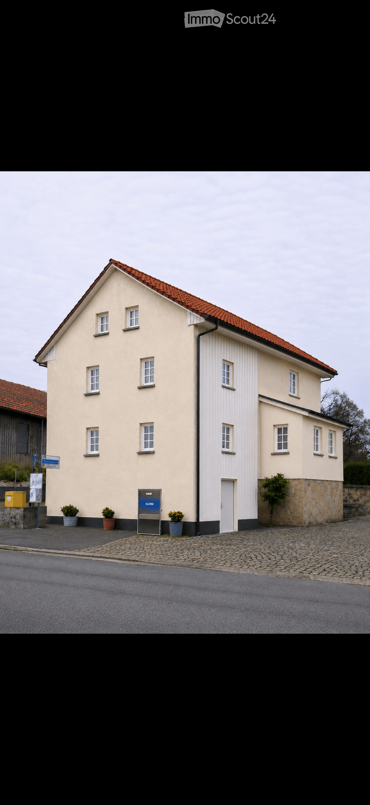 Two-story building with beige walls, red-tiled roof, multiple windows, and a white door. Potted plants and an alarm box are visible near the entrance.