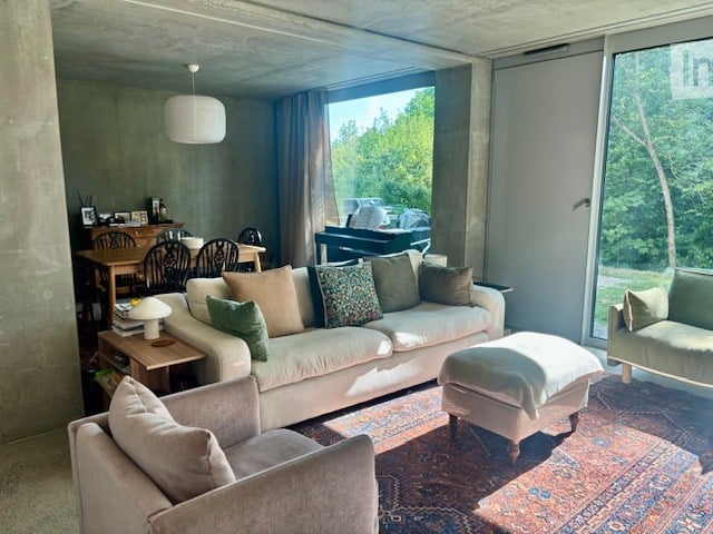 Modern living room with large windows offering a view of greenery, featuring a cream-colored sofa, ottoman, armchair, coffee table, dining table, and chairs.