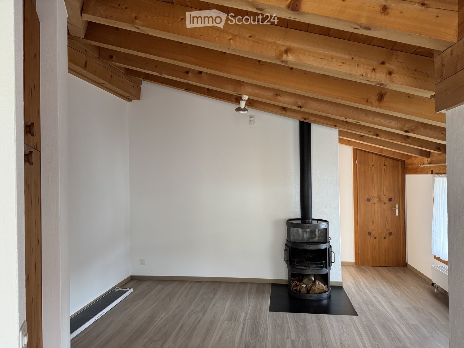 Empty room with wooden ceiling beams, light wooden flooring, white walls, and a black wood-burning stove with a chimney. A wooden door is visible in the background.