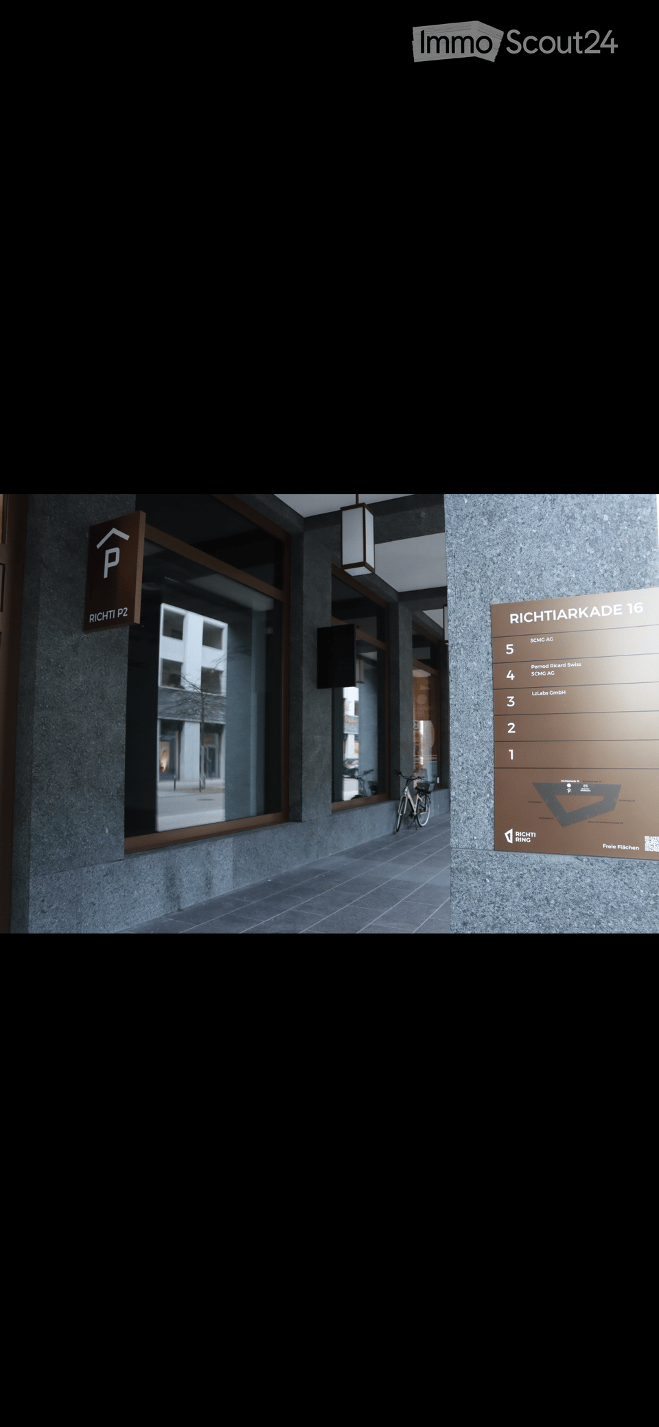 Modern hallway with a signboard listing office numbers, a bicycle parked nearby, and a view of a building through a large window.