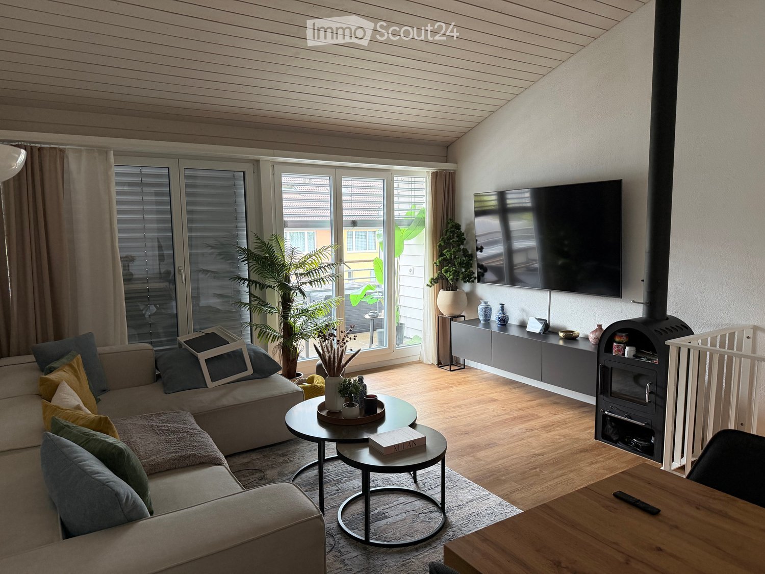 Modern living room with a grey sofa, coffee table, round side table, TV, fireplace, and decorative plants.