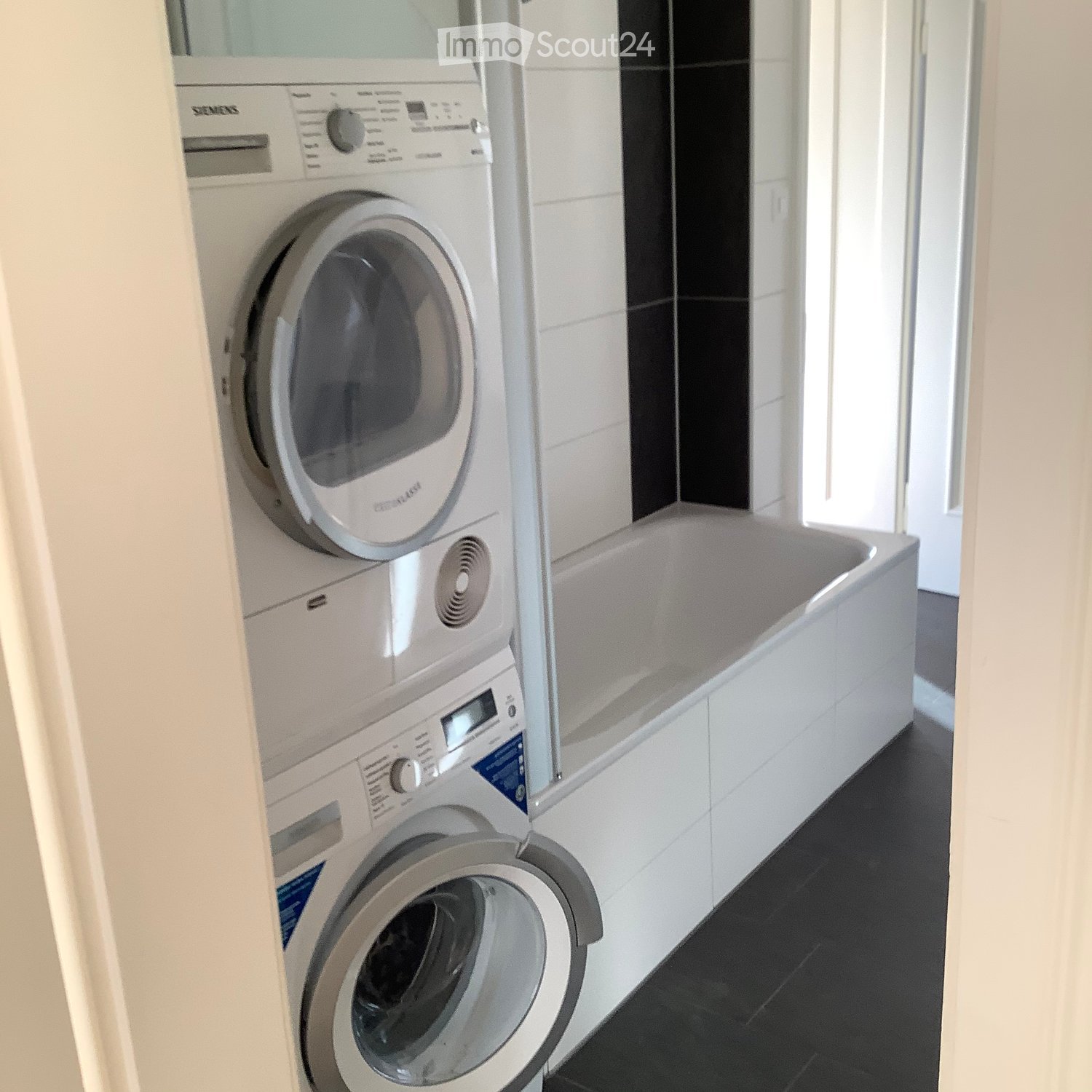 Modern bathroom with a bathtub, washing machine, and dishwasher, featuring white tiles and a black floor.