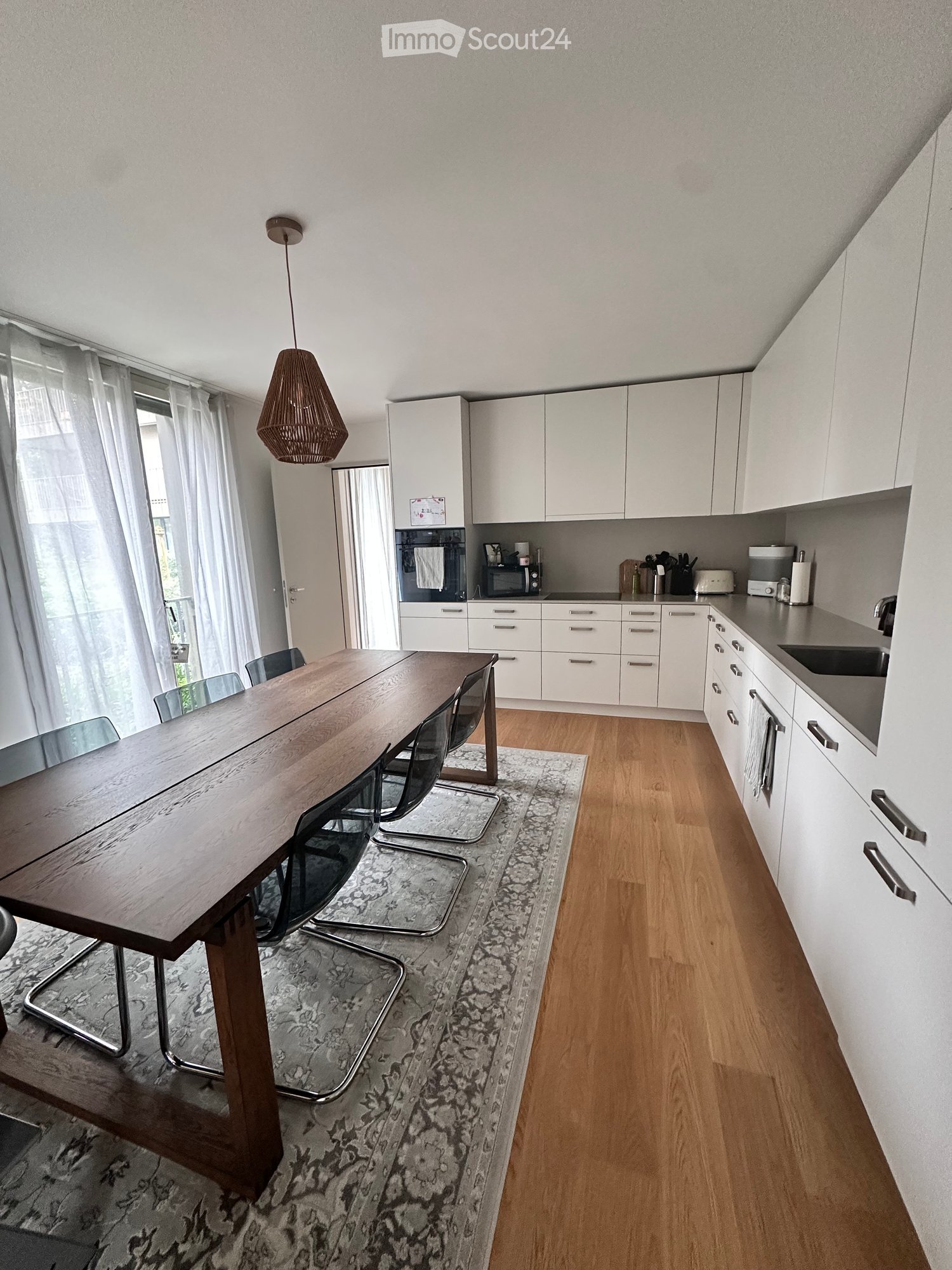 Modern kitchen with white cabinets, wooden floor, large wooden dining table with chairs, pendant light, and various appliances.