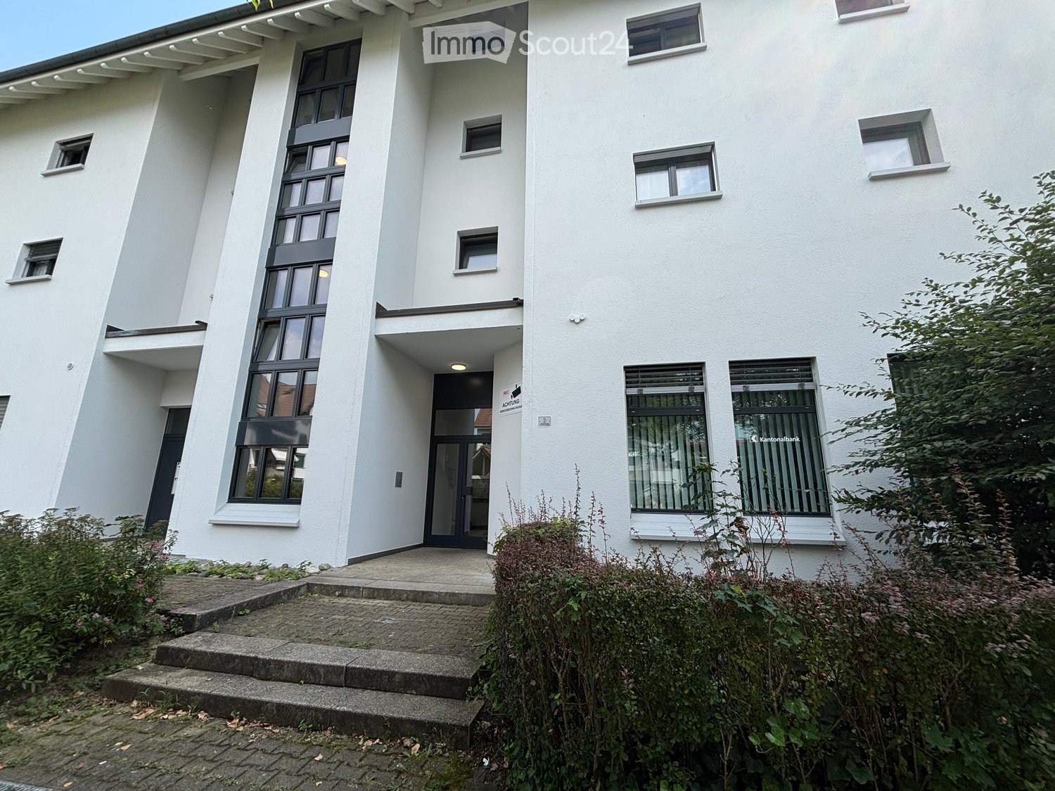 Modern white building with large windows, raised ground floor entrance, and surrounding greenery.