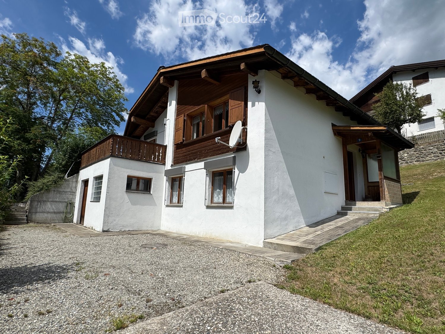 Two-story white house with wooden accents, satellite dish, and balcony on the upper level. The house is surrounded by greenery and has a gravel driveway.