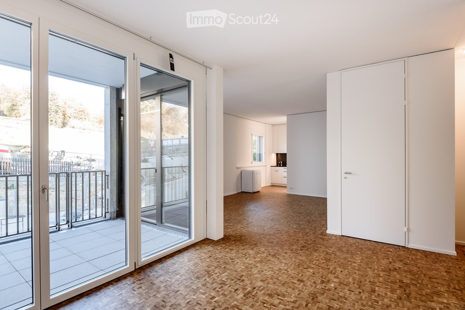 Empty room with wooden flooring, large sliding glass doors leading to a balcony, and a small kitchen area visible in the background.