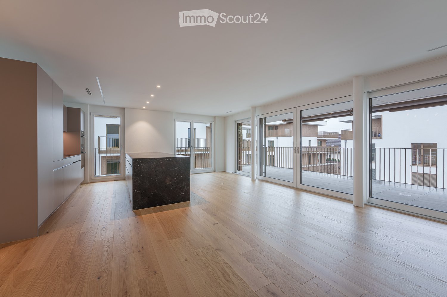 Bright, open-plan room with wooden flooring, large glass sliding doors leading to a balcony, and modern built-in kitchen units on one side.