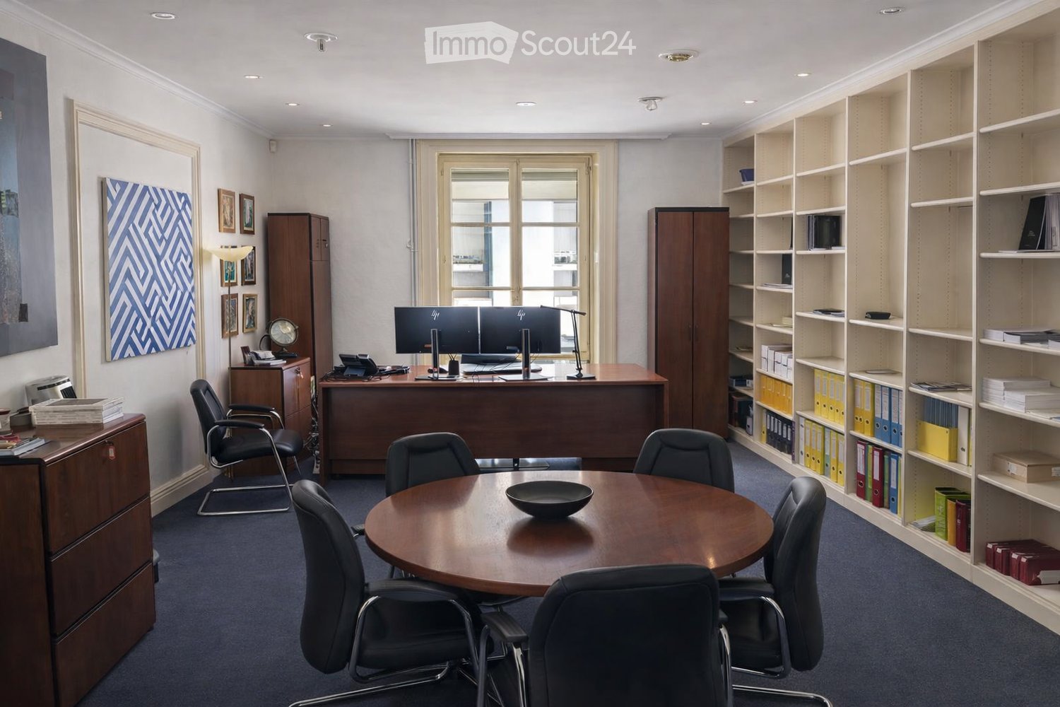 Office space with a large wooden desk, two computer monitors, a round meeting table with chairs, shelves filled with binders and books, filing cabinets, and artwork on the walls.