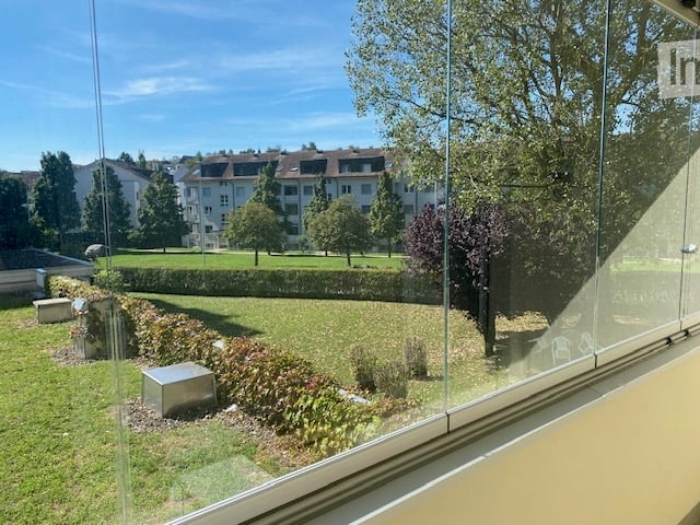 View from a room showing a well-maintained garden with grass, trees, shrubs, and a low hedge. The background includes a residential area with apartment buildings and parked cars.