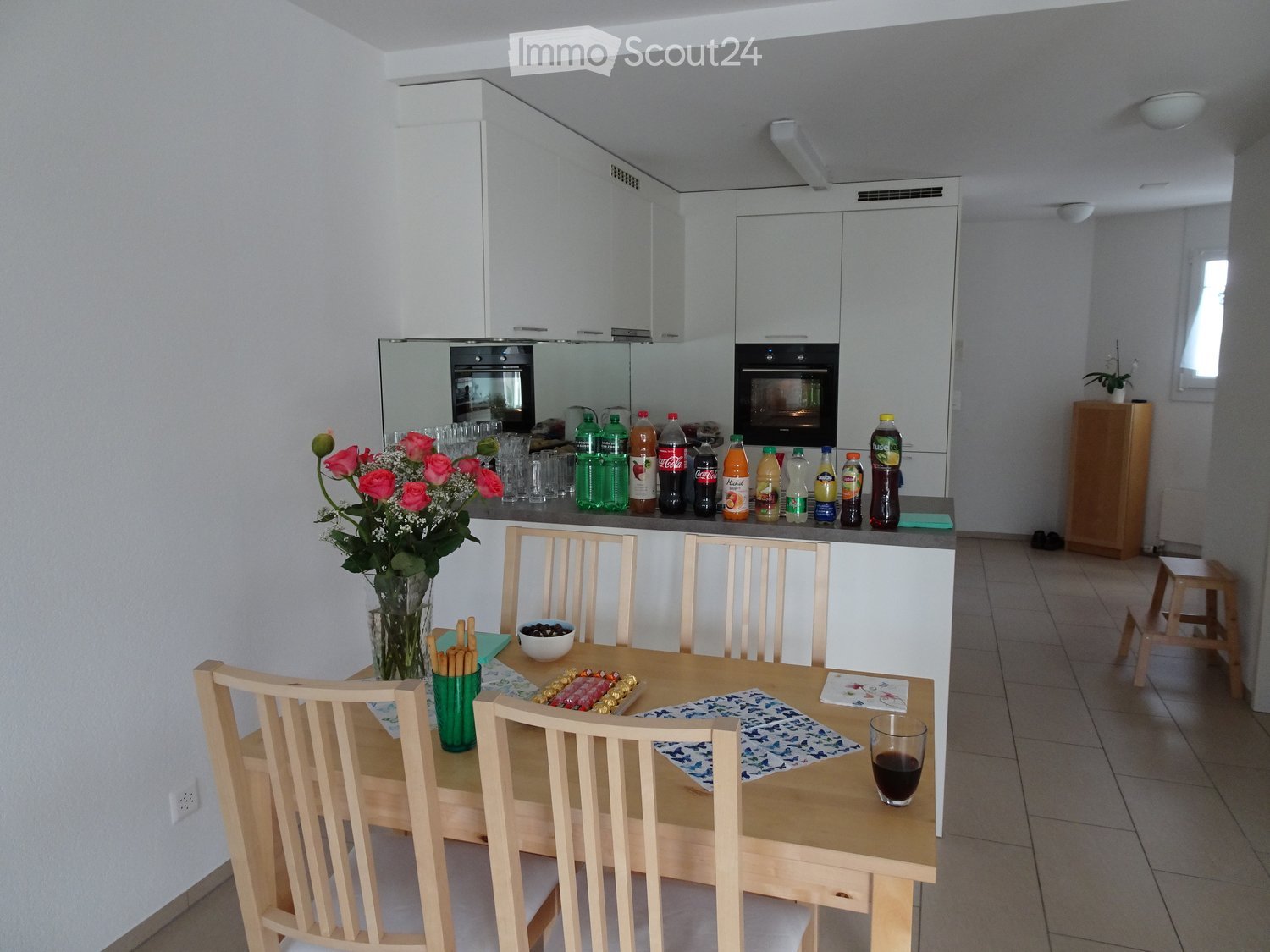 Modern kitchen with white cabinets, a wooden dining table with chairs, a vase of flowers, and various bottles on the counter.