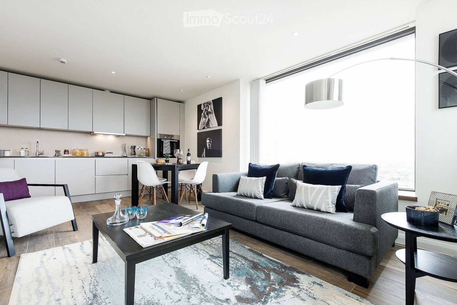 Modern living room with grey sectional sofa, coffee table, white dining chairs, and a sleek kitchen area with cabinets and a sink.