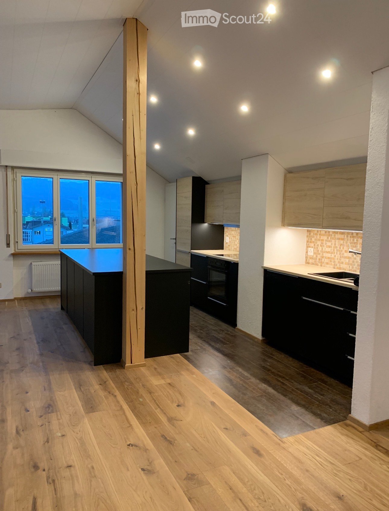 Modern kitchen with wooden flooring, black and light wood cabinets, a central island, and large windows overlooking a cityscape.