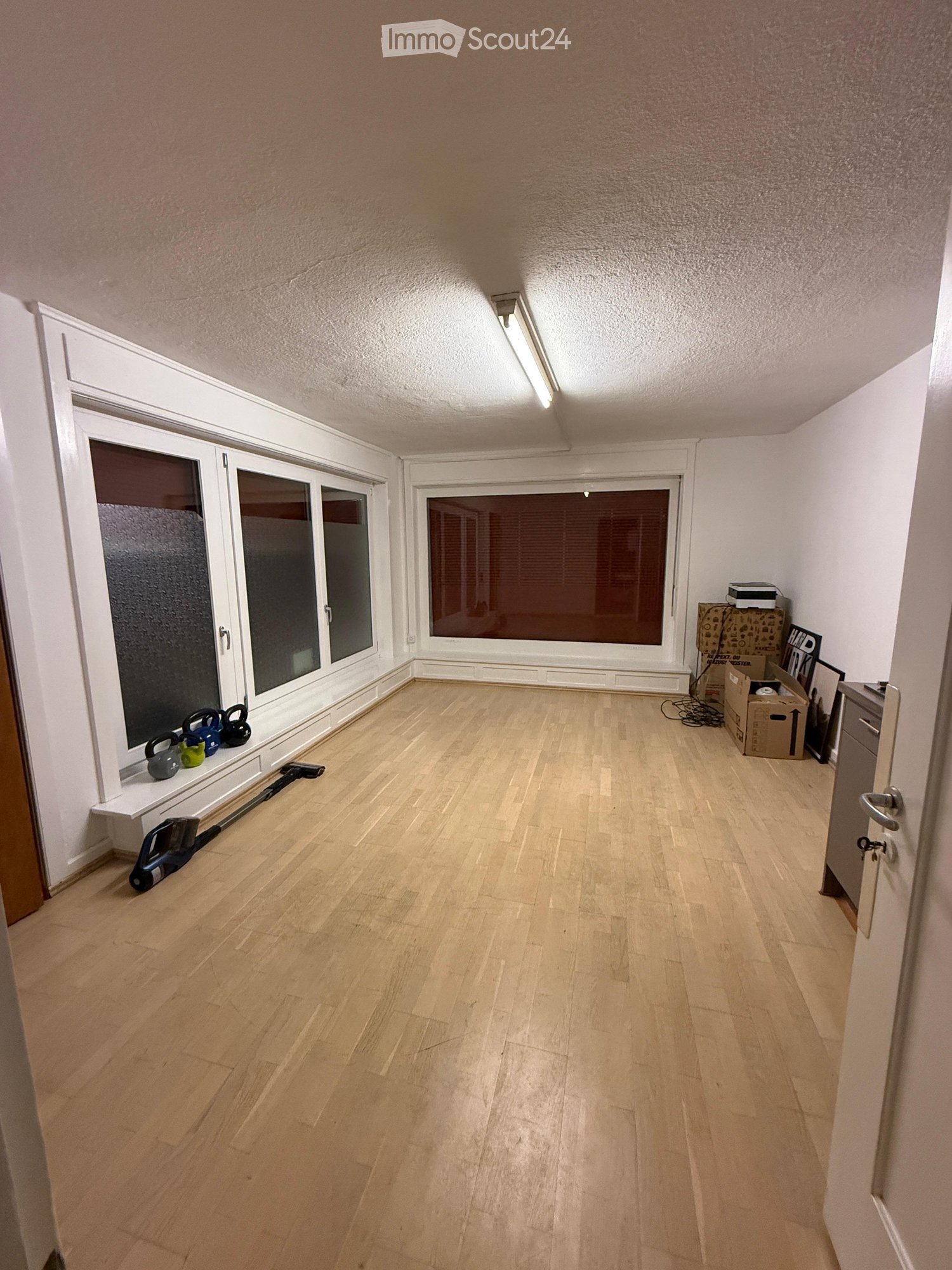 Empty room with light wooden flooring, white walls, large windows, and a door. No furniture or equipment visible.