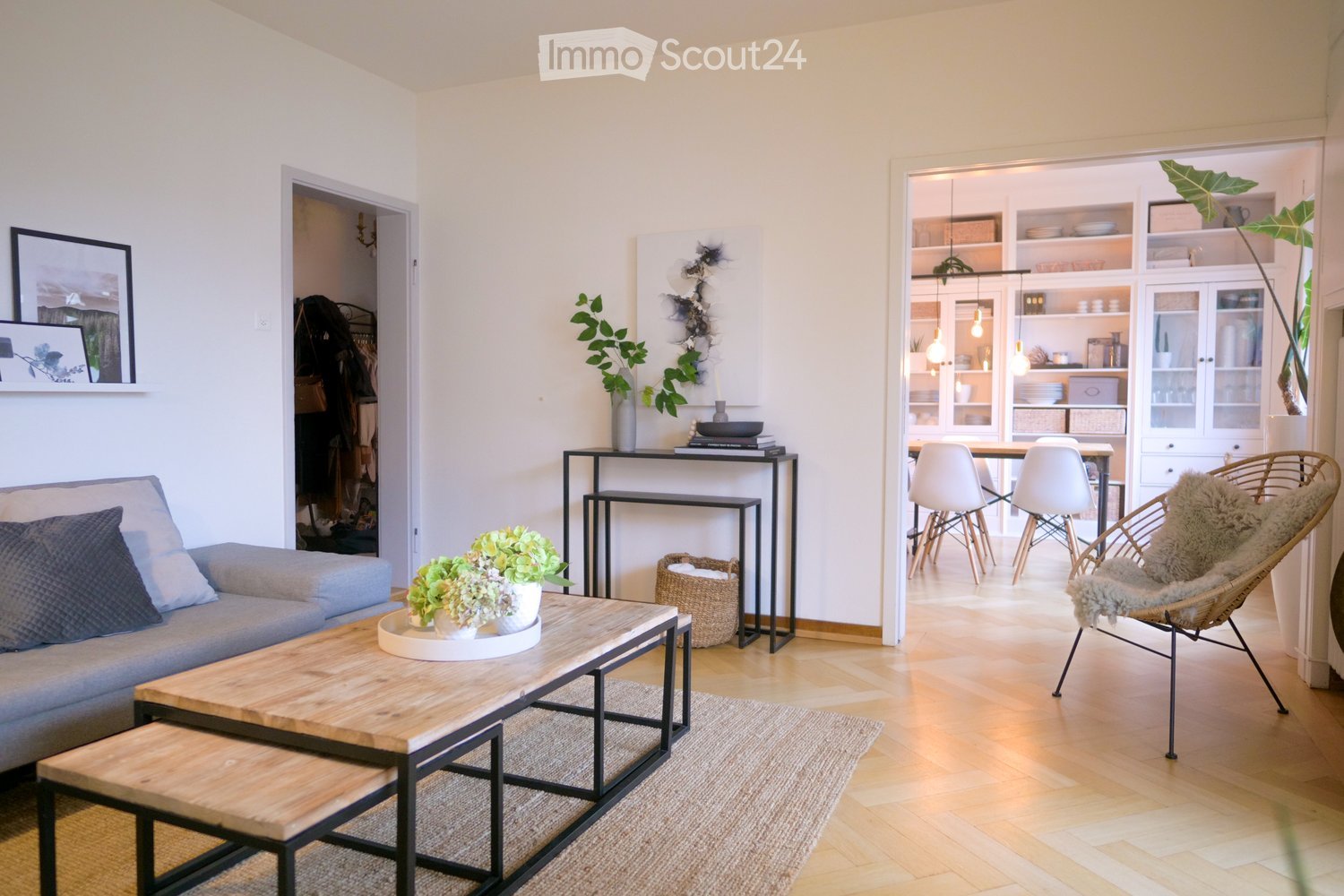 Modern living room with light wooden flooring, grey sofa, wooden coffee table, side table, dining area with white chairs and table, and decorative plants.